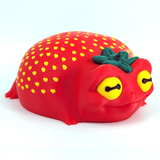 Strawg, the strawberry frog, sits on a white backdrop. He has a yellow eyes and seeds, a leaf hat, and a content expression on his face.