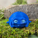 Front-facing view of Blueberry Frog with a soft smile and gentle expression, surrounded by moss and stone.