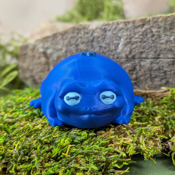 Front-facing view of Blueberry Frog with a soft smile and gentle expression, surrounded by moss and stone.