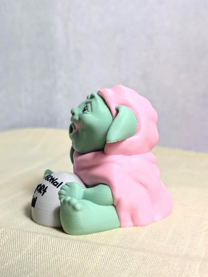 Angled view of the Emotional Support Goblin™ sitting with sign visible, resting calmly on a neutral surface.