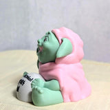 Angled view of the Emotional Support Goblin™ sitting with sign visible, resting calmly on a neutral surface.
