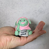 Green goblin figurine with a pink hood, held in a hand and holding a sign that says “It’s okay to rest.”