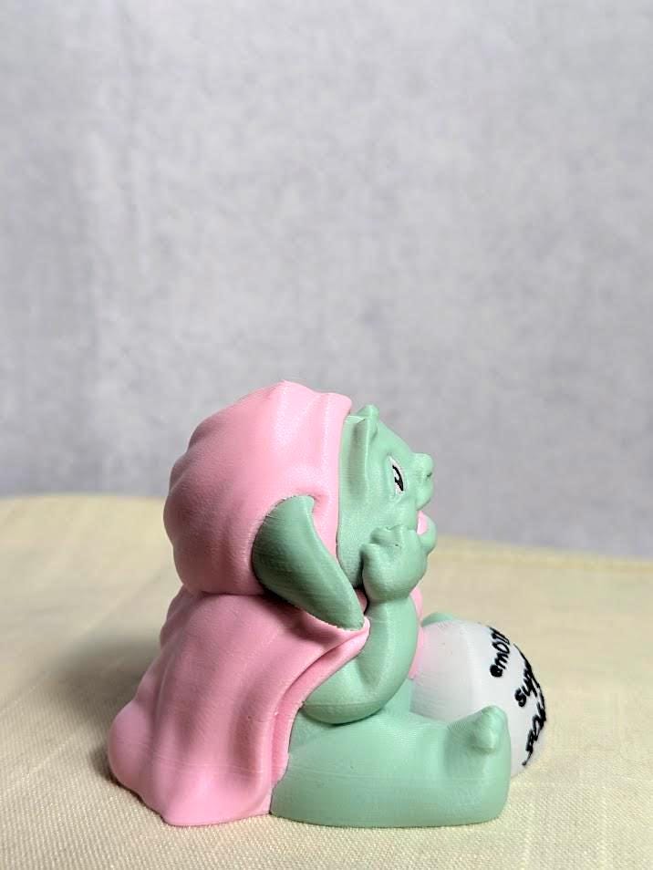 Side view of Emotional Support Goblin™ sitting cross-legged in a pink cloak, looking peaceful.
