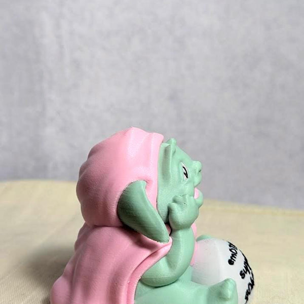 Side view of Emotional Support Goblin™ sitting cross-legged in a pink cloak, looking peaceful.