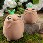 Two potato figurines posed on mossy ground in front of flowers, one in the foreground and one farther back for depth.
