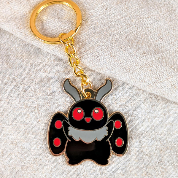 Cute Mothman enamel keychain with red eyes and gold chain on neutral fabric background