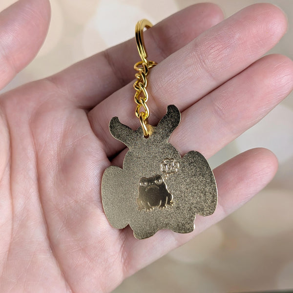 Reverse side of Mothman keychain held in hand showing engraved frog and gold finish