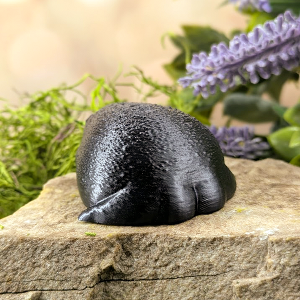 Rear view of a chonky black rainfrog figurine with textured skin, sitting on a stone surface surrounded by moss and flowers.