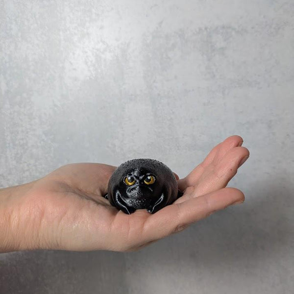 Small black frog figurine with angry yellow eyes, sitting in a person's palm like a furious goblin snack.