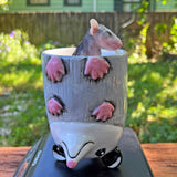 Small baby possum peeking out from a possum-themed ceramic cup on a wooden surface with a garden background.