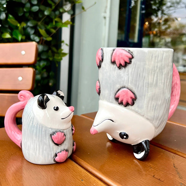 Two ceramic possum mugs with pink paws on a wooden surface.