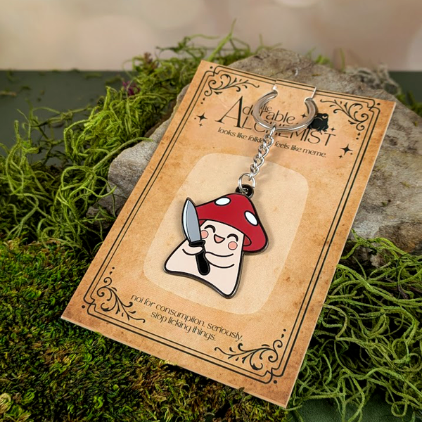 Keychain with mushroom design on a card, placed on a natural background with moss and stones.