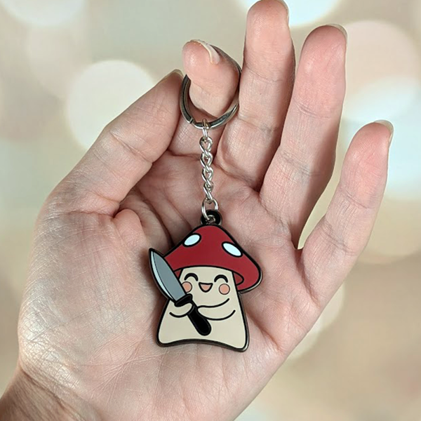 Keychain shaped like a mushroom with a face and knife held in a hand against a blurred background