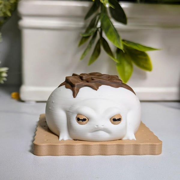 Front-facing close-up of a frog figurine styled like a s’more, with big eyes and a chocolate accent on its back, sitting neatly on a graham base.