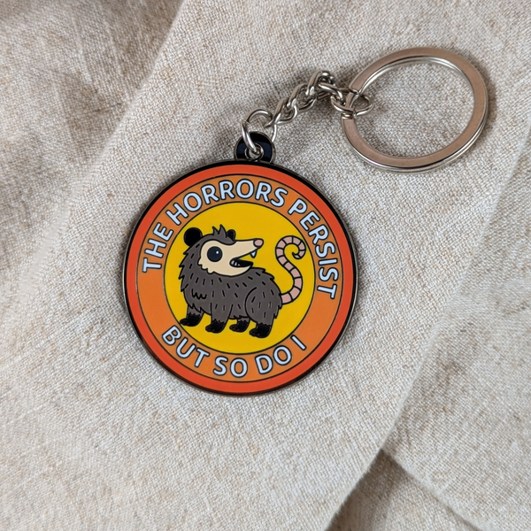 Funny possum enamel keychain with “The Horrors Persist But So Do I” text on beige background