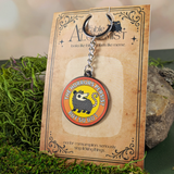 Keychain with possum design on a 'The Adorable Alchemist' branded card, placed on a natural background.