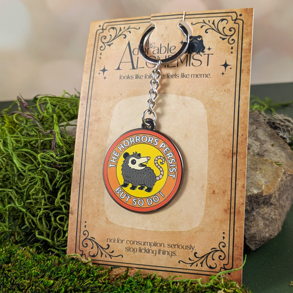 Keychain with possum design on a 'The Adorable Alchemist' branded card, placed on a natural background.