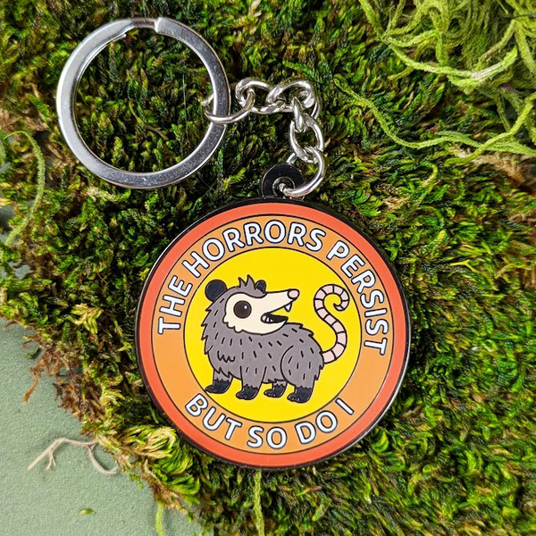 Keychain with an opossum and text 'The Horrors Persist But So Do I' on a moss background